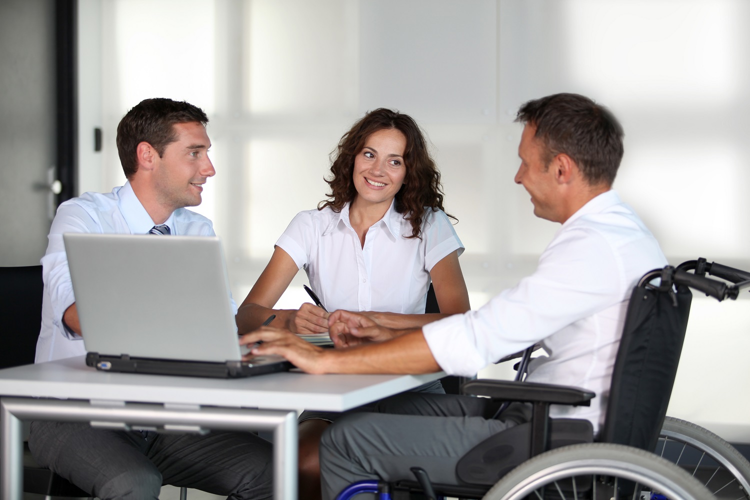 Ein Mann im Rollstuhl sitzt mit einem Mann und einer Frau am Tisch in einem Büro.